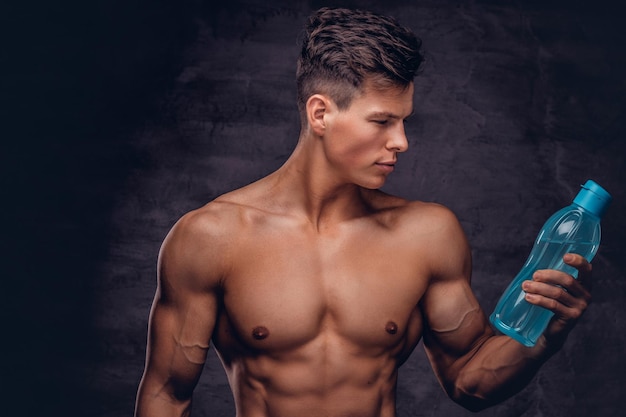 Portrait d'un modèle sexy de jeune homme torse nu avec un corps musclé et une coupe de cheveux élégante tient une bouteille d'eau rafraîchissante dans un studio. Isolé sur un fond sombre.