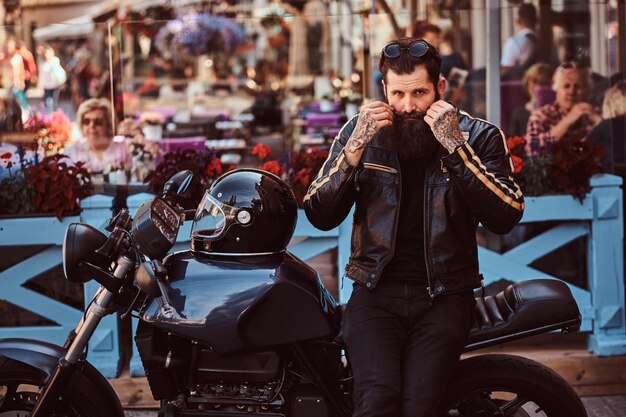 Portrait d'un motard à la mode élégant vêtu d'une veste en cuir noir, fixez la moustache tout en étant assis sur sa moto rétro sur mesure près de la terrasse d'un café.
