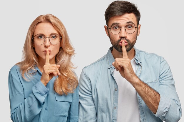 Portrait de muette jolie jeune femme et homme fait signe de silence, demandez à être calme, gardez les doigts sur les lèvres, tenez-vous étroitement, isolé sur un mur blanc. Concept de personnes, de complot et de secret