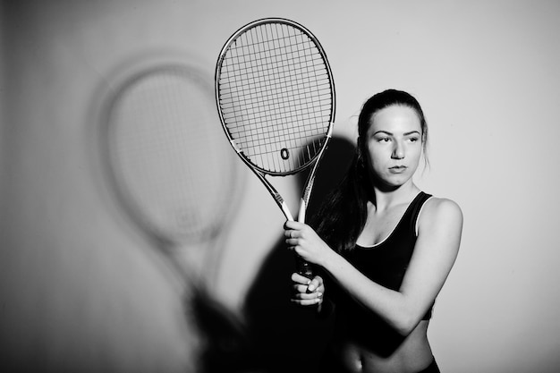 Portrait noir et blanc de la belle jeune femme joueuse en vêtements de sport tenant une raquette de tennis en se tenant debout sur fond blanc