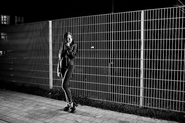 Portrait de nuit d'une fille modèle portant un jean et une veste en cuir contre une clôture en fer