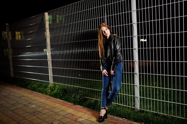 Portrait de nuit d'une fille modèle portant un jean et une veste en cuir contre une clôture en fer