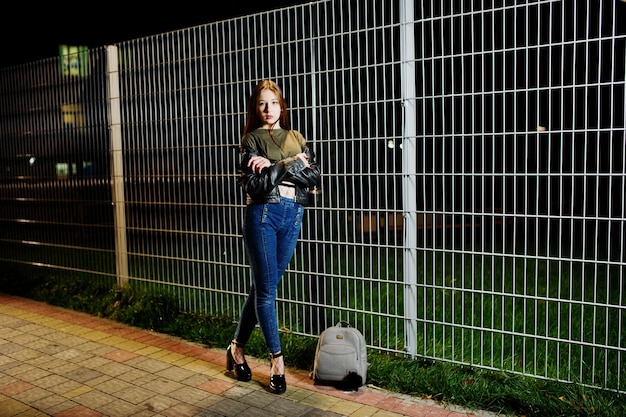 Portrait de nuit d'une fille modèle portant un jean et une veste en cuir contre une clôture en fer