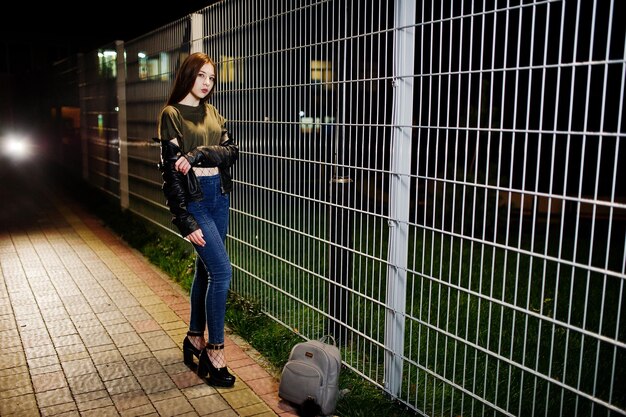 Portrait de nuit d'une fille modèle portant un jean et une veste en cuir contre une clôture en fer