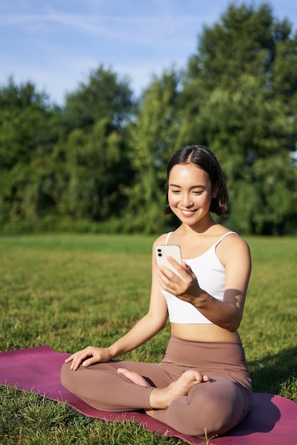 Portrait of asian girl sitting on mat in park parlant au smartphone méditant en ligne avec yoga inst