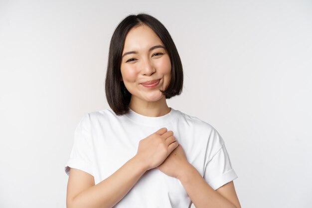 Portrait of carig young asian woman holding hands on heart regardant avec soin et amour touché par quelque chose se sentir flatté debout sur fond blanc