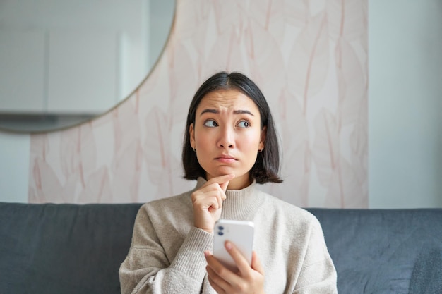 Portrait of Girl sitting on sofa with smartphone à la recherche réfléchie et hésitante au téléphone mobile scr