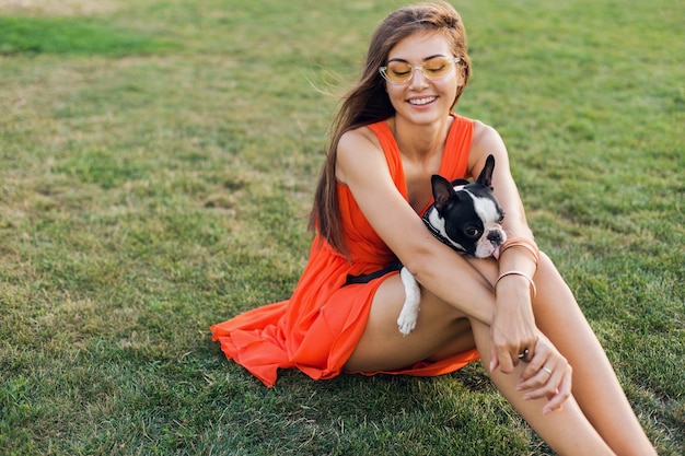 Portrait of happy pretty woman sitting on grass in summer park, holding boston terrier dog, smiling humeur positive, vêtue d'une robe orange, style branché, lunettes de soleil, jouant avec animal de compagnie