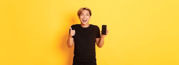 Portrait of happy smiling asian guy montrant l'écran du smartphone et thumbsup satisfait de l'application