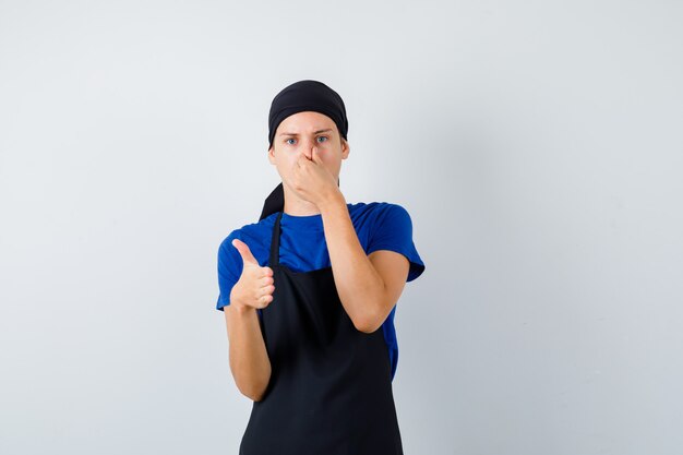 Portrait of male teen cook pinçant le nez et offrant une poignée de main en t-shirt, tablier et à la vue de face mécontent