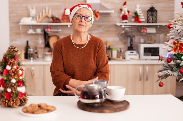Portrait of senior woman with santa hat standing at table dans la cuisine décorée de Noël