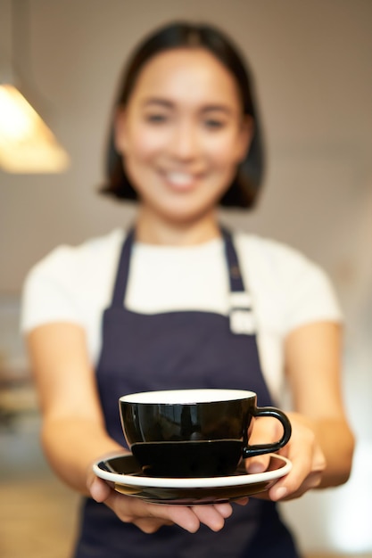 Photo gratuite portrait of smiling asian girl serveuse barista en uniforme de café vous donnant une tasse de café préparer dri
