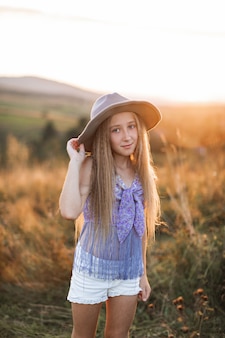 Portrait Of Smiling Little Girl With Long Blonde Hair Wearing Brown Hat, Standing In Summer Field Photo Premium