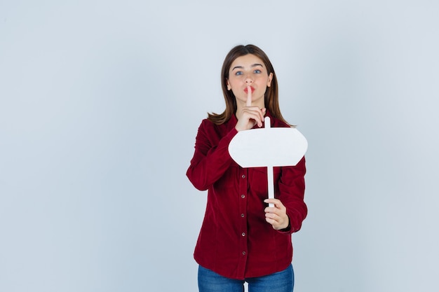 Portrait of teenage girl holding paper sign, montrant un geste de silence en chemise bordeaux et à la vue de face sensible