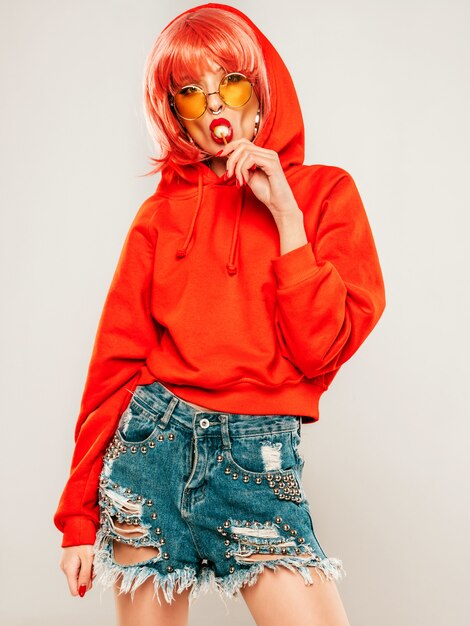 Portrait of young beautiful hipster bad girl in trendy red summer hoodie and earring in her nose.Sexy insouciante smiling blonde woman smiling in studio in wig.Positive model licking round round candy candy