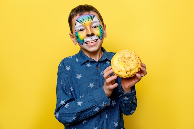 Photo gratuite portrait oh petit garçon en chemise décontractée avec de la peinture sur le visage, tenant un beignet sucré isolé sur un mur jaune.