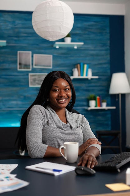 Portrait d'une personne joyeuse assise au bureau avec un ordinateur, travaillant à domicile sur un projet d'entreprise. Jeune femme regardant la caméra et utilisant un moniteur avec clavier pour faire du travail à distance.