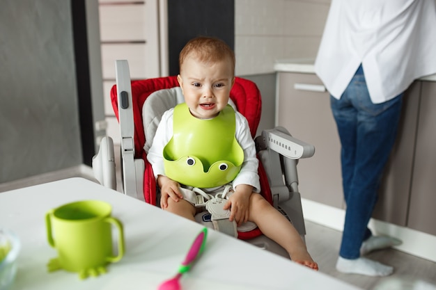 Portrait De Petit Garçon Effrayé Assis Dans Une Chaise De Bébé Dans La Cuisine, Pleurant Et Hurlant Pendant Que Sa Mère Lui Cuit La Nourriture.