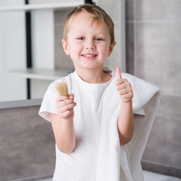 Portrait d&#39;un petit garçon heureux avec une serviette blanche sur son épaule, tenant le blaireau dans la main, montrant le pouce vers le haut de signe