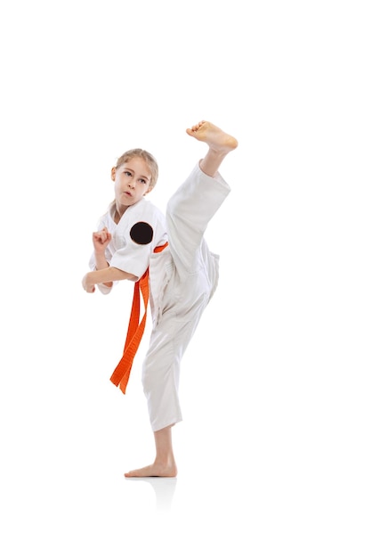 Photo gratuite portrait de petite fille, enfant en karaté de formation kimono isolé sur blanc. coup de pied latéral