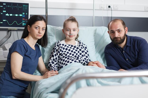 Portrait d'une petite fille malade se reposant dans un lit de clinique pédiatrique avec des parents inquiets et inquiets à côté d'elle tout en regardant la caméra. Famille moderne et décontractée dans la salle de récupération des patients de la clinique pédiatrique.