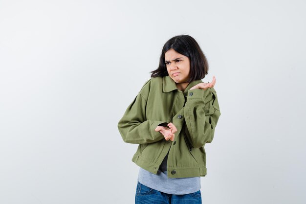 Portrait de petite fille montrant un geste impuissant en manteau, t-shirt, jeans et regardant la vue de face hésitante