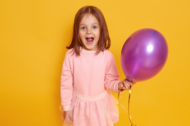 Portrait de petite fille surprise avec des cheveux raides foncés debout sur studio jaune de beaux vêtements, tenant un ballon violet dans les mains