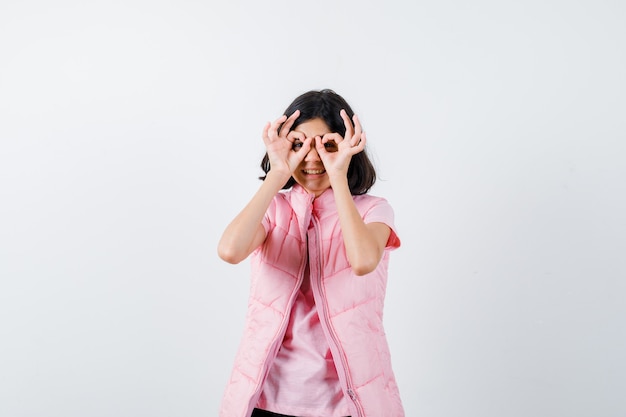 Portrait d'une petite fille en t-shirt blanc et gilet bouffant montrant le geste de lunettes