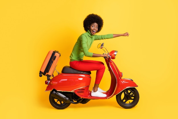 Portrait photo d'une femme afro-américaine brune excitée sur une moto avec un poing de bras de valise orange en avant isolé sur fond de couleur jaune vif