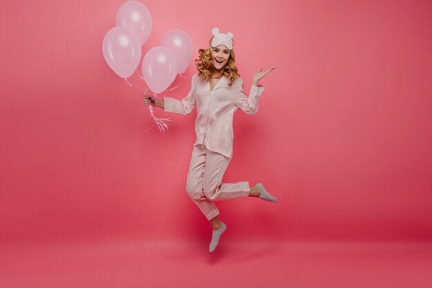 Portrait en pied d'agréable anniversaire fille en chaussettes sautant sur un mur rose. Jolie jeune femme en pyjama et masque de sommeil s'amusant avec des ballons à l'hélium.