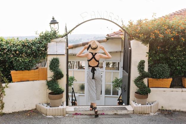 Portrait en pied de l'arrière de la femme au chapeau et vêtements rayés debout avec les jambes croisées devant les portes de fer noir