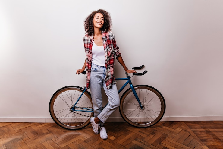 Photo gratuit portrait en pied d'une cycliste féminine attrayante. photo de studio d'une fille africaine intéressée en jeans debout sur blanc avec vélo.