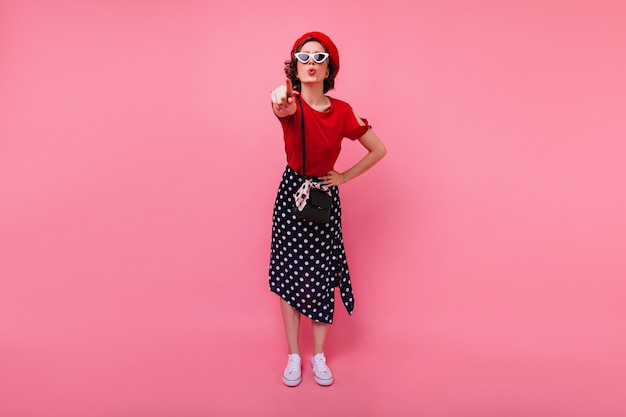 Portrait en pied de drôle de fille française pointant le doigt. magnifique femme caucasienne en jupe longue debout sur le mur rose.