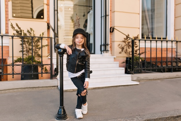 Portrait en pied de l'élégant enfant debout avec les jambes croisées à côté du pilier de fer en face de la boutique.