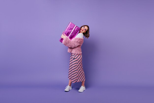 Portrait en pied d'une femme caucasienne heureuse en baskets tenant la boîte présente. Tir à l'intérieur d'un modèle féminin élégant posant avec un cadeau d'anniversaire et souriant.