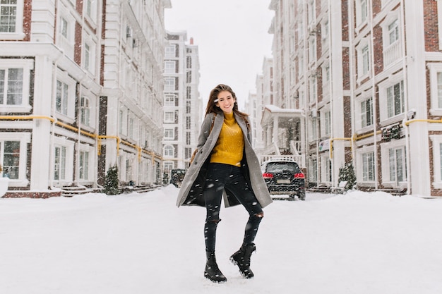 Portrait en pied d'une femme gracieuse porte un chandail jaune à la mode qui s'amuse dans la rue sous les chutes de neige. Photo extérieure d'une femme européenne mince en manteau chaud dansant sur la rue urbaine.