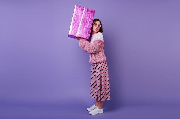 Portrait en pied d'une femme intéressée en baskets blanches tenant une grande boîte cadeau. fille d'anniversaire glamour.