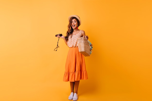 Portrait en pied d'une femme sensuelle porte des chaussures blanches et un chapeau de paille. Fille caucasienne bien habillée faisant des grimaces sur orange.