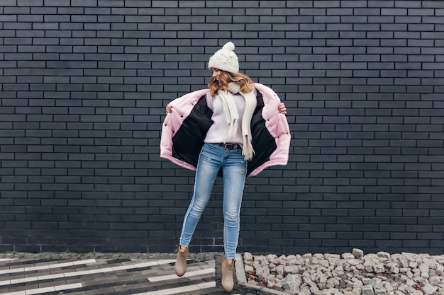 Portrait en pied d'une fille mince en jeans et veste rose dansant dans la rue. Tir extérieur d'un magnifique modèle féminin en bonnet tricoté exprimant des émotions positives.