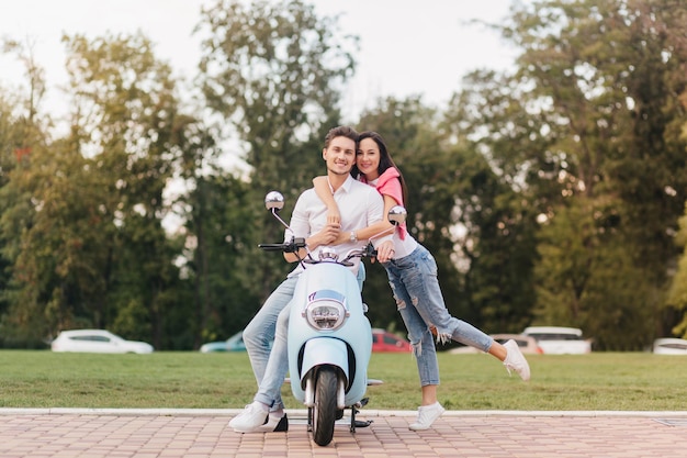 Portrait en pied d'une fille à la mode en jeans posant avec un mec heureux assis sur un scooter. Portrait en plein air d'une jeune femme inspirée passant du temps dans le parc avec son petit ami.