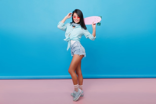 Portrait en pied d'une fille sportive porte de jolies chaussettes et une chemise en coton élégante. Slim femme latine brune avec planche à roulettes bénéficiant dans la chambre avec intérieur bleu.