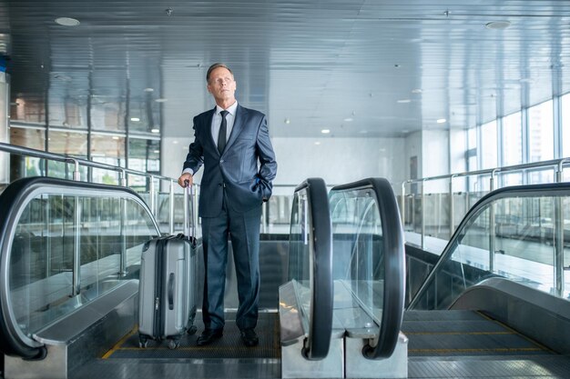 Portrait en pied d'un homme pensif sérieux avec la valise trolley descendant de l'escalator