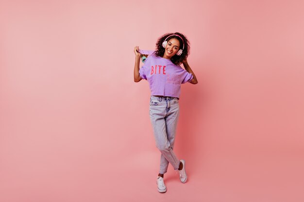 Portrait en pied d'une incroyable dame africaine en jeans tenant une planche à roulettes. Élégante fille noire dans les écouteurs et chemise violette debout sur le rose.