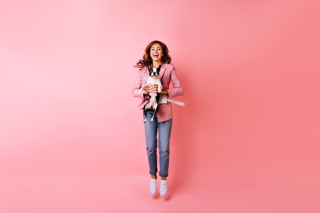 Portrait en pied de jeune femme extatique sautant avec bouledogue français. Portrait intérieur d'une fille rousse émotionnelle se détendre avec son animal de compagnie.