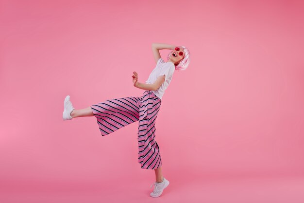 Portrait en pied de jeune femme en pantalon à la mode dansant avec un sourire heureux. Tir intérieur d'une fille élégante mince en perruque rose s'amusant