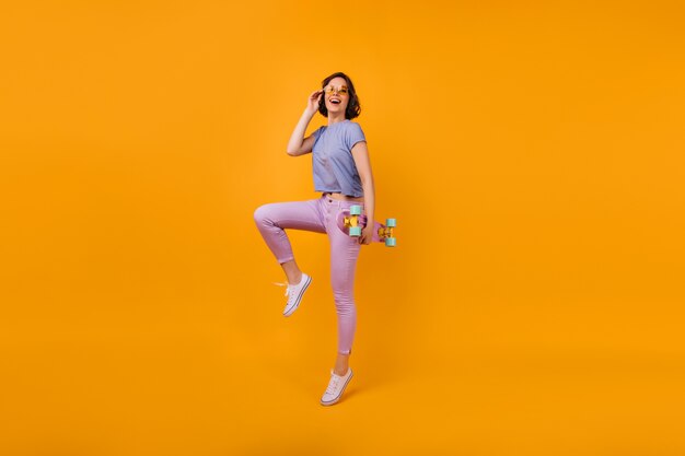 Portrait en pied d'une jolie femme aux cheveux courts avec longboard. Plan intérieur d'une fille spectaculaire en tenue d'été et lunettes de couleur jaune.