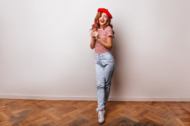 Portrait en pied de jolie femme aux cheveux longs en béret français. Belle fille au gingembre posant avec sucette.