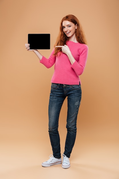 Portrait en pied d'une jolie fille rousse satisfaite, pointant le doigt sur une tablette numérique