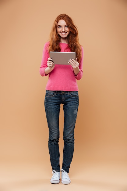 Portrait en pied d'une jolie fille rousse souriante tenant une tablette