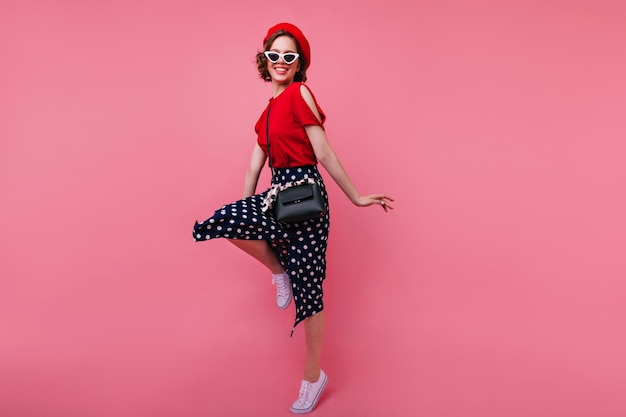 Photo gratuite portrait en pied d'un modèle féminin de bonne humeur en béret rouge français dansant avec le sourire incroyable fille bouclée en lunettes noires sautant en studio